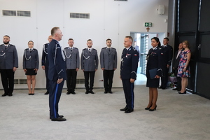 na zdjęciu na pierwszym planie widoczni są umundurowani policjanci, jeden stojący przed komendantem i komendant na drugim planie widoczni sa umundurowani policjanci stojący w dwóch rzędach