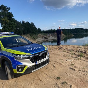 na zdjęciu widoczny jest oznakowany radiowóz o0raz umundurowany policjant  w oddali zbiornik wodny