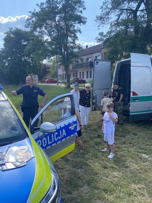 ma zdjęciu widoczny jest oznakowany radiowóz ,, umundurowany policjant  dzieci koło  niego