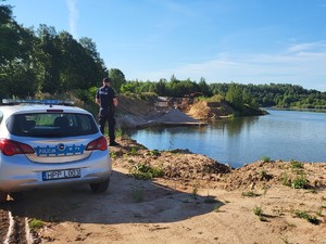 na zdjęciu widoczny  jest umundurowany policjant oznakowany radiowóz , policjant stoi na zbiornikiem wodnym obok radiowozu