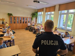 na zdjęciu widoczny  jest policjant tyłem w mundurze i widoczne są dzieci siedzące w ławkach