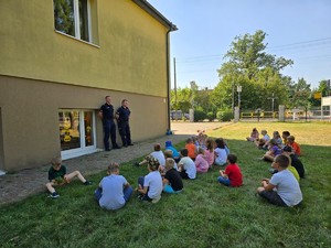 na zdjęciu widocznych jest dwóch stojących umundurowanych policjantów i dzieci siedzące na trawie