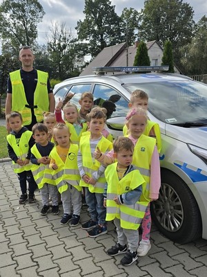 na zdjęciu widoczna jest grupa dzieci w kamizelkach odblaskowych stojąca obok oznakowanego radiowozu wśród dzieci stoi umundurowany policjant w kamizelce odblaskowej