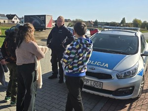 na zdjęciu widoczny jest oznakowany radiowóz umundurowany policjant i dzieci stojące obok niego