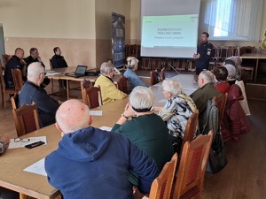 na zdjęciu widoczny jest umundurowany policjant osoby starsze siedzące wokół stołu w tle widoczny jest ekran na którym wyświetlana jest prezentacja dotycząca bezpieczeństwa osób starszych