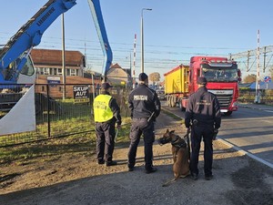 na zdjęciu widoczni są umundurowani funkcjonariusze Straży Ochrony Kolei umundurowany policjant siedzący obok pies z założonym kagańcem. Mundurowi stoją w pobliżu przejazdu kolejowego.