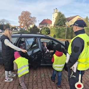 na zdjęciu widoczny jest umundurowany policjant dwoje dzieci w kamizelkach odblaskowych kobieta wszyscy stoją pry samochodzie osobowym w którym otwarte są tylne drzwi