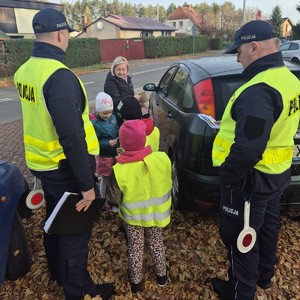 na zdjęciu widoczni są dwaj umundurowani policjanci w kamizelkach odblaskowych pośrodku nich stoi dziecko w kamizelce odblaskowej kobiet oraz dziecko bez kamizelki w tle widoczny jest samochód osobowy