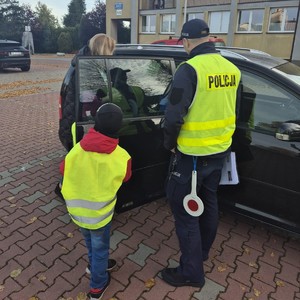 na zdjęciu widoczny jest umundurowany policjant oraz dziecko kamizelce odblaskowej , policjant również ma kamizelkę odblaskową