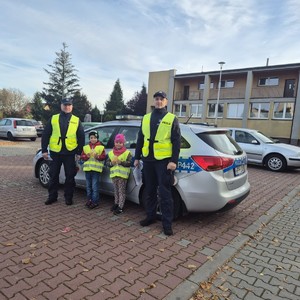 na zdjęciu widocznych jest dwóch umundurowanych policjantów w kamizelkach odblaskowych między nimi stoi dwoje dzieci w kamizelkach odblaskowych za nimi widoczny jest oznakowany radiowóz