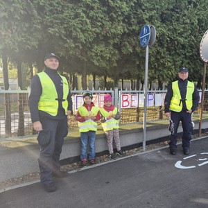 na zdjęciu widocznych jest dwóch u mundurowanych policjantów oraz dwoje dzieci za nimi widoczne jest metalowe ogrodzenie na którym wisi transparent z napisem jedz bezpiecznie w fotelikach