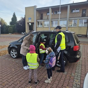 na zdjęciu widoczny jest umundurowany policjant dwoje dzieci , jedno z nich ma kamizelkę widać również kobietę stojąca przy samochodzie osobowym w którym otwarte są tylne drzwi