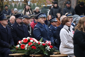 na zdjęciu widoczni są ludzie siedzący na ławce na cmentarzu wojskowym oraz umundurowani policjanci w galowych mundurach widoczny jest również wieniec z kwiatów.