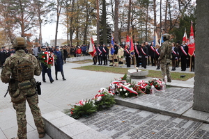 na zdjęciu widoczni są policjant i policjantka w galowych mundurach niosący wieniec w kierunku pomnika , widoczny jest również umundurowany żołnierz stojący do nich przodem