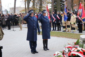 na zdjęciu widoczny jest policjant i policjantka w mundurze galowym , którzy stoją przed pomnikiem , oboje salutują przed nimi leżą wieńce