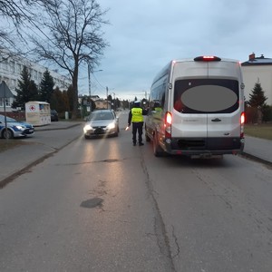 na zdjęciu widoczny jest umundurowany policjant w kamizelce odblaskowej stojący na środku drogi pomiędzy dwoma pojazdami busem i samochodem osobowym