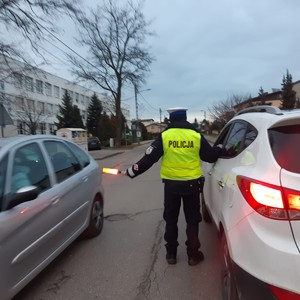 na zdjęciu widoczny jest umundurowany policjant w kamizelce odblaskowej stojący na środku drogi pomiędzy dwoma pojazdami osobowym