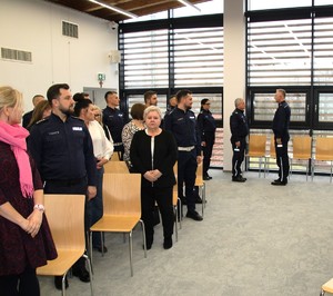 na zdjęciu widoczna jest duża sala na której poustawiane są krzesła przy nich umundurowani policjanci jak również osoby cywilne w tle stoi dwóch umundurowanych policjantów na wprost siebie