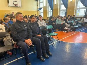 na zdjęciu widocznych jest dwoje policjantów w mundurach siedzą na krzesłach  na sali gimnastycznej za nimi siedzą dzieci