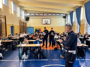 na zdjęciu widoczne są dzieci siedzące przy ławkach ustawionych na sali gimnastycznej widoczny jest również umundurowany policjant