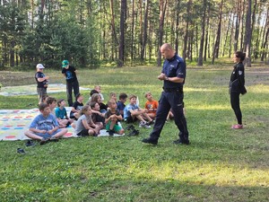 na zdjęciu widoczny jest umundurowany policjant przed nim na kocu siedzi grupa dzieci obok stoi kobieta
