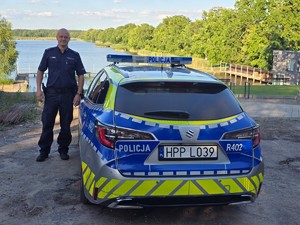 na zdjęciu widoczny jest umundurowany policjant stojący obok oznakowanego radiowozu za jego plecami widoczny jest zbiornik wodny
