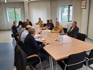 na zdjęciu widoczny jest stół wokół którego siedzą umundurowali strażacy i policjanci oraz osoby w cywilnych ubraniach