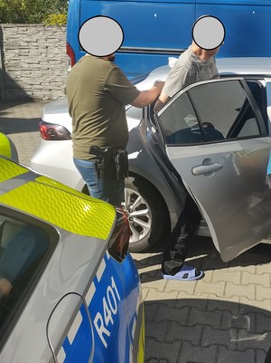 na zdjęciu widoczny jest policjant w cywilnym ubraniu , który pomaga wsiąść do nieoznakowanego radiowozu zatrzymanemu mężczyźnie widoczna jest również część stojącego obok radiowozu oznakowanego