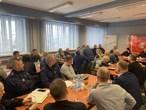 na zdjęciu widoczne są osoby mężczyźni i kobiety siedzące przy stołach na dużej sali wśród nich są umundurowany funkcjonariusze wojska straży miejskiej straży ochrony kolei. Widoczny jest sztandar Komendy Powiatowej Policji w Lublińcu jak również siedzący przy stole umundurowani policjanci.