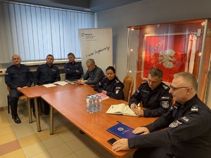 na zdjęciu widoczni są umundurowani policjanci siedzący przy stole z a ich plecami widoczna jest szklana gablota w której znajduje się sztandar Komendy Powiatowej Policji w Lublińcu