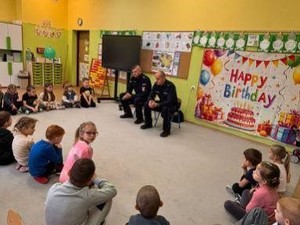 na zdjęciu widocznych jest dwóch umundurowanych policjantów a w koło nich siedzące na dywanie dzieci w  wieku przedszkolnym