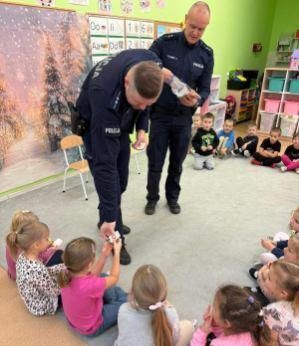 na zdjęciu widocznych jest dwóch umundurowanych policjantów stojących i rozdających odblaski  siedzącym na dywanie dzieciom w  wieku przedszkolnym