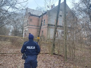 na zdjęciu widoczny jest umundurowany policjant stojący na wprost nieotynkowanego domu pustostanu