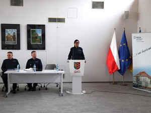 na zdjęciu widoczna jest umundurowana policjantka stojąca za mównica obok przy stole siedzi dwóch umundurowanych policjantów w tle widoczna jest flaga polski i unii europejskiej