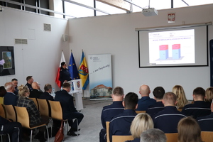 na zdjęciu widoczna jest duża sala na której widoczni są zaproszeni goście siedzący na krzesłach na ścianach wiszą obrazy , widoczny jest stół za którym siedzą umundurowani policjanci obok przy mównicy stoi umundurowana policjantka wśród siedzących gości widoczni są również umundurowani policjanci