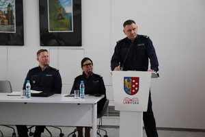 na zdjęciu widoczna jest umundurowana policjantka i umundurowany policjant siedzący przy stole obok przy mównicy stoi umundurowany policjant