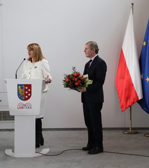 na zdjęciu widoczna jest kobieta stojąca za mównicą obok niej stoi mężczyzna z bukietem kwiatów w ręce