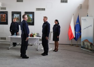 na zdjęciu widoczni są policjanci i policjantka stojący na baczność w tle widoczne są flagi Polski i Unii Europejskiej