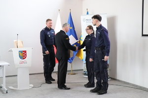 na zdjęciu widoczni są umundurowani policjanci i policjantka jak również umundurowany strażak w tle widoczna jest mównica strażak wręcza pismo policjantom