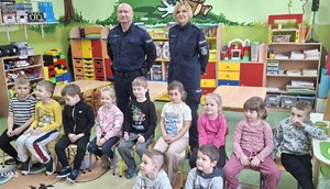 na zdjęciu widoczne są dzieci w wieku przedszkolnym siedzące w sali za ich plecami stoją umundurowani policjanci