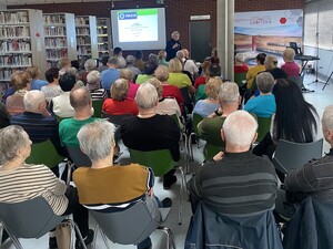 na zdjęciu widoczna jest duża sala na której siedzą ludzie na krzesłach przed nimi stoi umundurowana policjantka za jej plecami widoczny jest ekran z boku widoczne saąregały z książkami