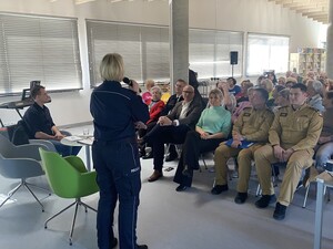 na zdjęciu widoczna jest stojąca policjanta z mikrofonem obok siedzi nieumundurowany policjant przed policjantka w pierwszym rzędzie siedzą umundurowali strażacy strażnik straży miejskiej oraz kobieta w cywilnych ubraniach