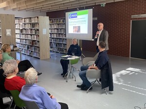 na zdjęciu widoczny jest mężczyzna z mikrofonem obok siedzi na krześle przy stoliku policjantka a z drugiej strony policjant w cywilnym ubraniu