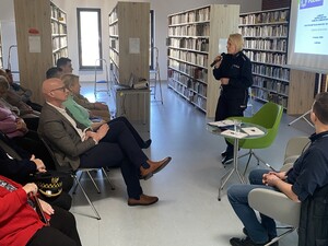 na zdjęciu widoczna jest policjantka trzymająca mikrofon przed nią siedzą ludzie za nią widoczny jest ekran