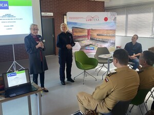 na zdjęciu widoczna jest kobieta trzymająca mikrofon obok stoi policjanta za nimi widoczny jest duży ekran