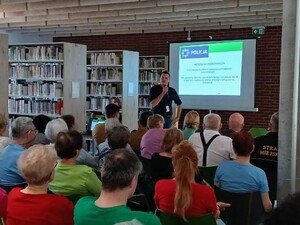 na zdjęciu widoczny jest policjant w cywilnym ubraniu trzyma mikrofon za nim wyświetlany jest slajd na ekranie przed nim siedzą ludzie