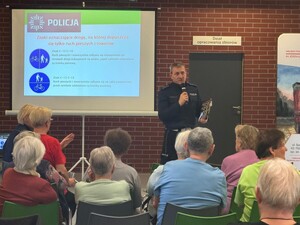 na zdjęciu widoczny jest policjant z ruchu drogowego przed nim siedzą ludzie z anim jest ekran na którym wyświetlany jest slajd