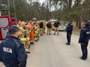 policjanci strażacy stoją w rzędzie przed nimi dwoje policjantów za nimi wozy strażackie