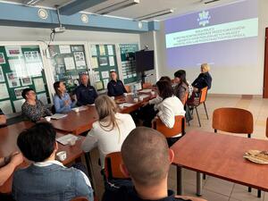 na dużej sali przy stole siedzą kobiety wśród nich siedzą umundurowani policjanci jeden policjant stoi widać również duży ekran