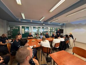 na dużej sali przy stole siedzą kobiety wśród nich siedzą umundurowani policjanci jeden policjant stoi widać również duży ekran
